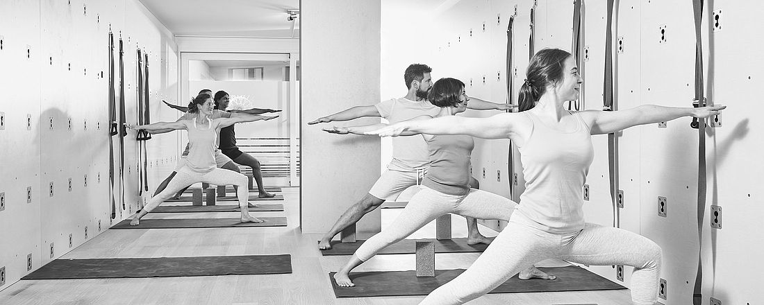 Ein Gruppe übt den Krieger 2 im Studio von the yoga place Zürich. Man sieht die Yogawand. 