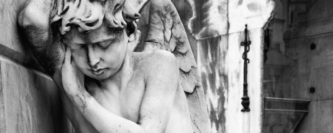 Schwarz weiss Bild: Ein Engel lehnt sich seitlich in Trauer an einer Wand. Er stützt den Kopf mit seinem rechten Arm.